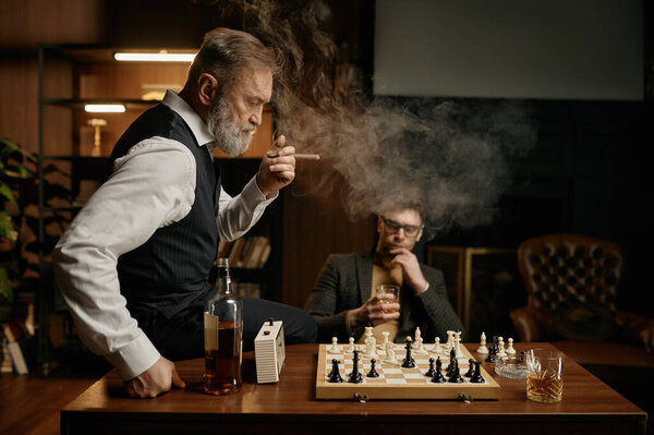 Two chess players taking break for smoking cigar and planning strategy of game. Selective focus on senior man looking at chessboard