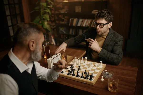 2人の男が自宅のリビングルームのテーブルに座ってチェスをしている シニアの父と大人の息子は 一緒にウイスキーや喫煙葉巻を飲みながら夜を過ごす インテリジェントゲーム — ストック写真