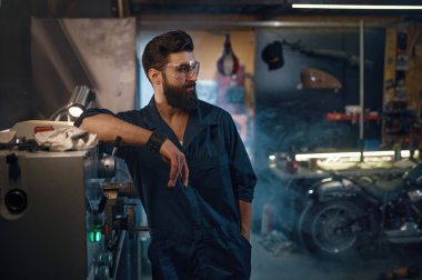 Modern motosiklet atölyesinde duran koruyucu gözlüklü ve tulumlu kendine güvenen yakışıklı tamircinin portresi. Motosiklet bakımı ve profesyonel servis konsepti