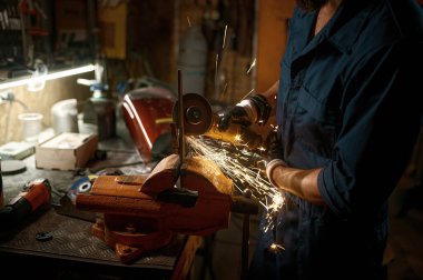 Tamirci atölyede çalışırken yedek parçayı elle yapıp monte ediyor. Teknisyen elinde kırpılmış görüş ve makineden çıkan kıvılcımlar