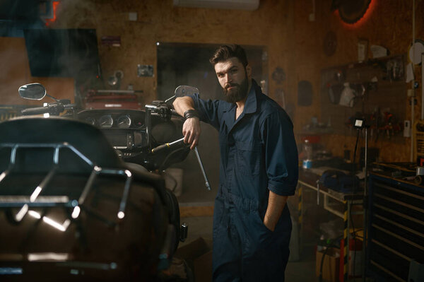 Handsome brutal repairman in work wear looking at camera portrait. Bearded young workman standing nearby motorcycle and holding wrench over smoky dark repair garage workplace
