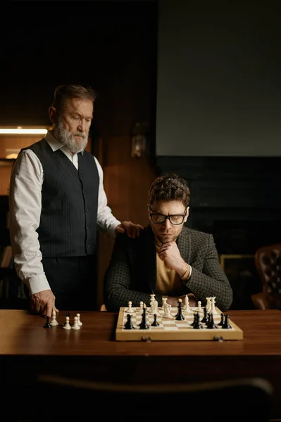 若い男がチェスボードを見て 高齢者の父親のヒントを聞く ホームライブラリで戦略的なゲームをプレイするシニア親の教師の息子 週末のインテリジェントなアクティビティコンセプト — ストック写真