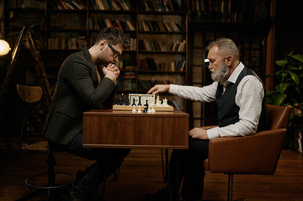 Concentrated chess players holding competition match at home library room. Senior man and young guy playing strategic game using stopwatch for measuring time