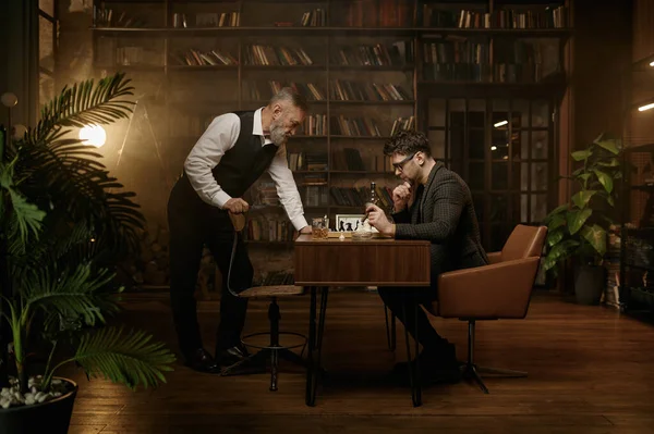 居心地の良い家庭のインテリアの上にチェスボードを見て2人の神経チェス選手 シニアと大人の紳士は面白いマッチ喫煙シガーと飲酒スコッチ — ストック写真