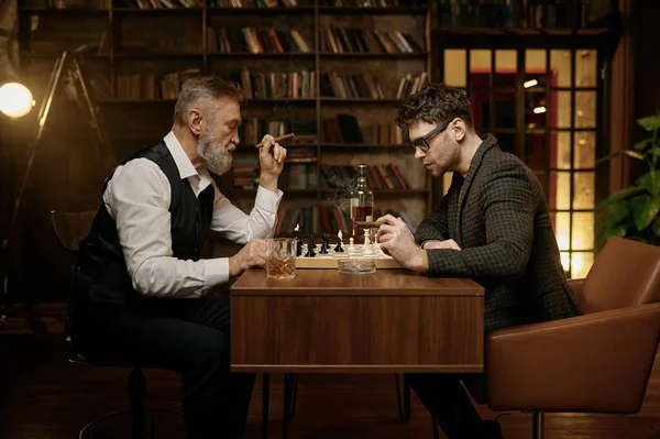 チェスをしたり 葉巻を吸ったり ウィスキーを飲んだりする聡明な人々の家族 シニアと若い男性がチェス盤を見てテーブルに座っている スマートゲーム — ストック写真