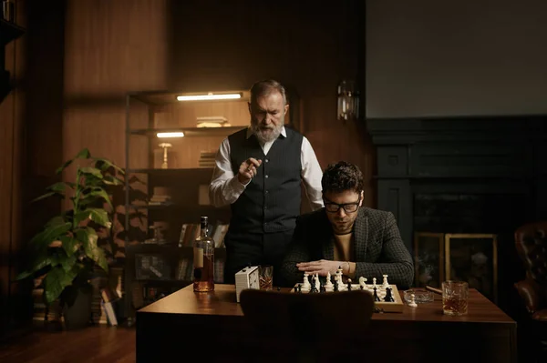 居心地の良い家庭のインテリアの上にチェスボードを見て2人の神経チェス選手 先輩と大人の紳士は面白いマッチ喫煙葉巻を持っています — ストック写真