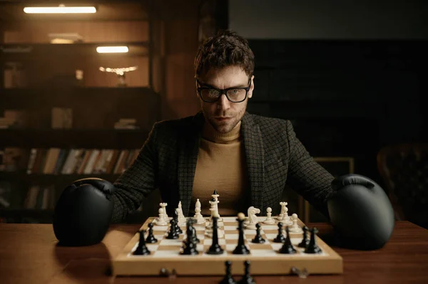 対戦相手と戦う準備ができているチェス盤とテーブルに座っているボクサー手袋を身に着けている怒っている若い男 肖像画撮影 — ストック写真