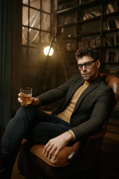 革のアームチェアに腰掛けながら ガラスからウイスキーやスコッチアルコールを飲む若い大人のエレガントな紳士の肖像画 — ストック写真