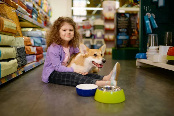 Little girl child sitting on pet shop floor holding corgi dog on knees. Shopping at supermarket offering goods for domestic animals