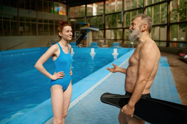 水泳プールで先輩男性と話す若い女性インストラクター アクアエアロビクストレーニングの前または後の古い男性水泳の質問に答える女性トレーナーに焦点を当てる — ストック写真