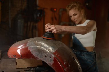 Motosiklet garajında ağır iş yapan genç bir kadın metal bir motoru tamir etmek için öğütücü kullanıyor.