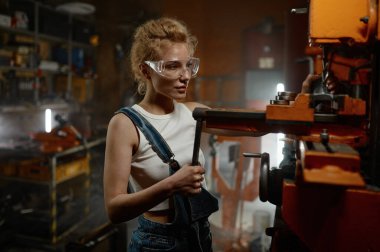 Garaj atölyesinde güvenlik koruyucu gözlük takan, elektrikli değirmen ve sondaj makinesi üzerinde çalışan profesyonel, ciddi genç bir kadının yakın görüntüsü.