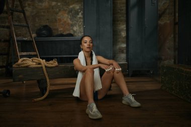 Kadın boksör boynunda havluyla yerde dinleniyor. Spor kıyafetli genç bir kız oturmuş eski spor salonunun iç tarafındaki kameraya bakıyor.