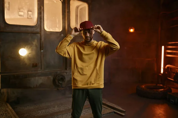 stock image Portrait of man brake dancer touching his cap. B-boy posing for camera at studio with loft interior design and train on background
