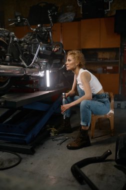 Düşünceli genç bir kadın elinde kilit anahtarı tutarak özel motorsikletle sorunu çözmeye çalışıyor. Akşamları atölye içi