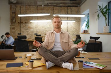 Sakin adam ofis çalışanı meditasyon yapıyor ve nefes alıyor. İş yerindeki masa başında lotus pozisyonunda oturuyor.