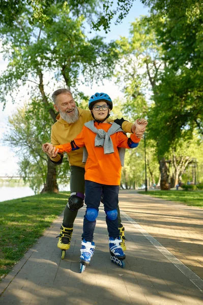 Elderly man enjoying sports in sunny summer park with his little son. Happy overjoyed father following boy riding roller skates