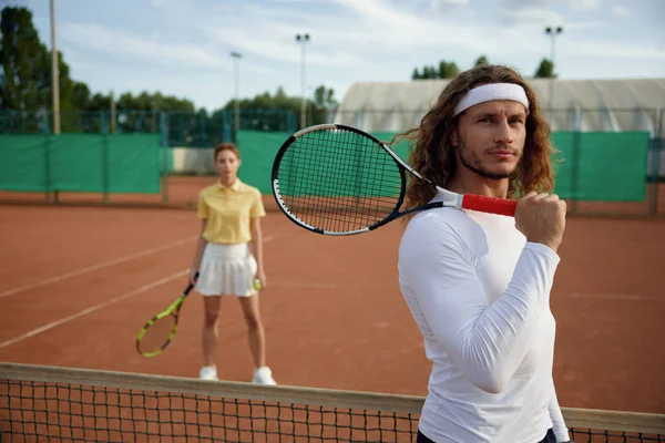 裁判所でテニスをする友人たち 肩にラケットを持ち カメラを見ている自信のある男性選手に焦点を当てます 若者カップルのための週末のスポーツレクリエーション — ストック写真