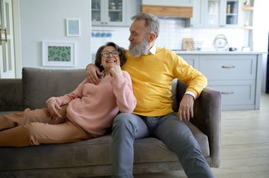 Mutlu yaşlı çift evde oturma odasında kanepede dinlenirken gülümsüyor ve sarılıyor.