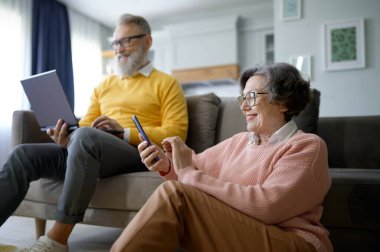 Mutlu emekli çift evdeki oturma odasında dinlenirken dijital cihaz kullanıyor. Yaşlı kadın akıllı telefondan arkadaşlarıyla sohbet ediyor ve kıdemli koca dizüstü bilgisayardan sosyal medyaya mesaj atıyor.