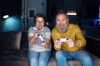 Üst düzey çift evde video oyunu oynuyor. Yaşlı karı koca kanepede oturup ellerinde joysticklerle hafta sonları boş vakit geçirmekten zevk alıyorlar.