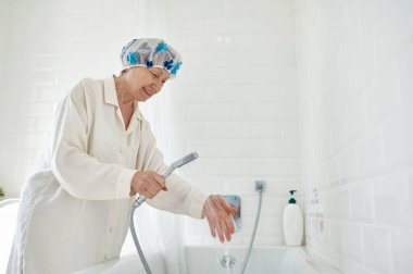 Bornozlu mutlu yaşlı kadın küveti suyla dolduruyor banyo yapmaya hazırlanıyor. Yurtiçi yaşam tarzı ve emeklilik konusunda günlük rutin