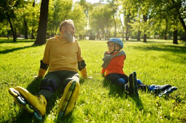 Neşeli adam ve çocuk patenciler parkta sergilenen çimlerin üzerinde dinleniyor. Genç ve yetişkin insanlar birlikte aktif boş zaman aktiviteleri ve eğlenceli spor zamanları geçiriyorlar.