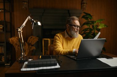 Üst düzey bir adam akşam evde çalışırken ve masada otururken dizüstü bilgisayarda klavye kullanıyor.