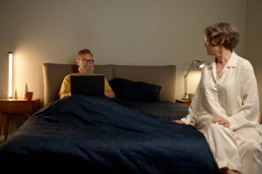 Mutlu emeklilik. Yatmadan önce yatak odasında yaşlı bir çift var. Mutlu bir eş, dizüstü bilgisayarla çalışan kocasına bakıyor.