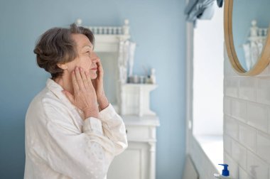 Yaşlı bir kadın banyoda aynanın önünde bornoz giyerek anti-yaş kremi sürüyor. Emekli kadın için günlük cilt bakımı ve hijyen rutini