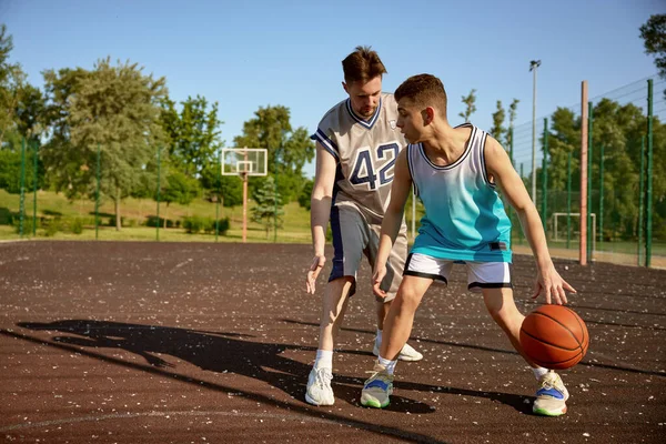 Young trainer teaching teenage boy playing basketball on street court. Sport class for teen boys