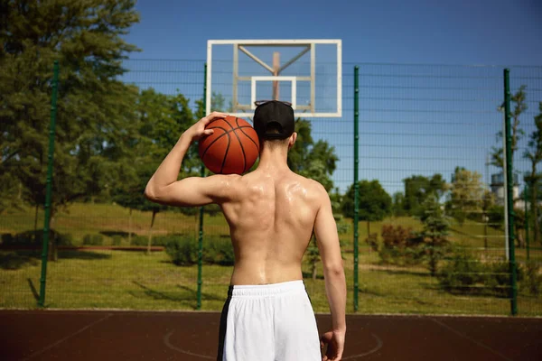 肩にボールを握っているシャツレスのバスケットボール選手の筋肉体の背中からの眺め 屋外スポーツトレーニング — ストック写真