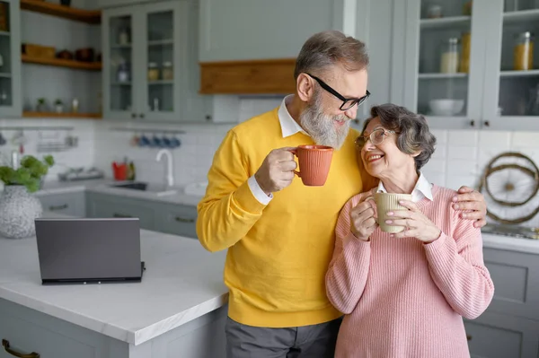 Happy senior couple drinking coffee or tea on home kitchen. Portrait of elderly husband and wife hugging and looking at each other enjoying retirement together