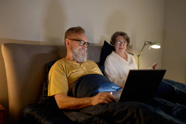 Senior couple using laptop computer while lying on bed in bedroom at comfortable home. Smiling husband showing interesting video to loving wife