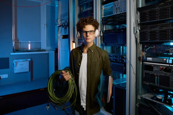 Smart teenager student having internship in modern data center. Male IT technician wearing headphones holding laptop computer carrying wires for solving problem with network connection