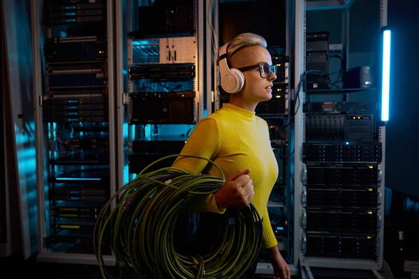 Young woman IT technician wearing headphones carrying wires bundle walking through server rack in big data center inside