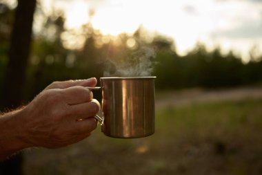 Yakın plan seçici odaklı buharı tüten yürüyüş fincanı erkek eliyle çok güzel doğal bir arka plan sunuyor. Seyahat macerası, yürüyüş eğlence aktivitesi konsepti.