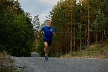 Güçlü emekli sporcu çam ormanları yolunda hızla koşuyor. Sporcu dışarıda koşarken kalori yakıyor.