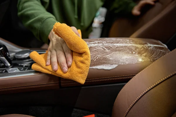 Closeup car detailing studio worker cleaning leather interior using foam detergent and tissue napkin
