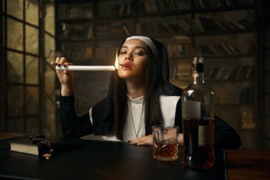 Ahlaksız Hıristiyan rahibenin yanan mumdan yanan sigarası masada açık viski şişesiyle oturuyor. Manastır kostümü giymiş ahlaksız bir kadının portresi.