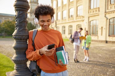 Tasasız, rahat, kulaklıklı genç bir çocuk dışarıda müzik dinlemek için cep telefonu kullanıyor. Lisede, üniversitede ya da üniversitede ara