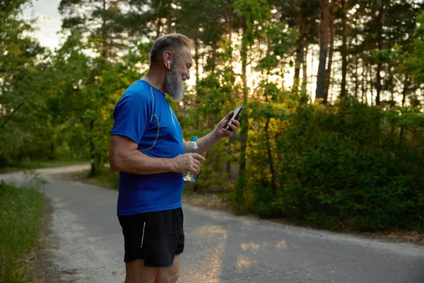 Retired man jogger taking break during training exercises outdoors drinking water watching funny video on phone