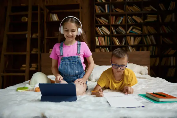 ハッピースクールの子供たちは 自宅で一緒に宿題を勉強し 準備しています 弟がコピーブックでタスクを書いている間 ヘッドセットとデジタルタブレットデバイスを使用してリスニング教師 — ストック写真