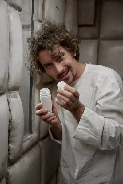 Crazy man with psychiatric disorder smiling ominously to camera holding opened pills bottle standing in hospital padded room
