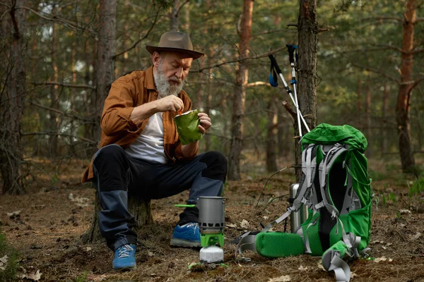 Relaxed senior man hiker eating food and taking break after touristic trip in forest. Outdoors adventure on retirement