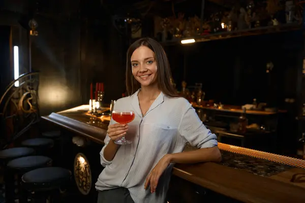 Portrait of relaxed cheerful young woman holding cocktail while sitting at bar counter looking at camera with smile