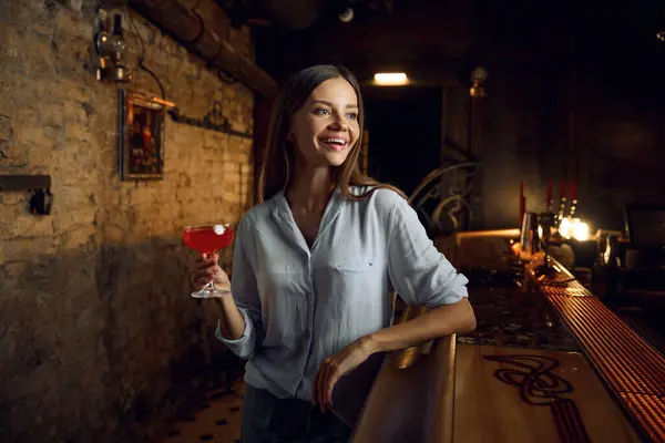 Portrait of relaxed cheerful young woman holding cocktail while sitting at bar counter and laughing