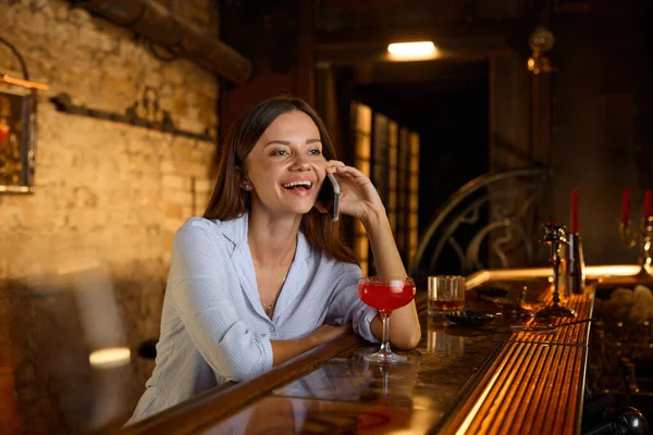 Happy smiling woman talking phone with friend while drinking cocktail and resting in bar. Leisure time activity in evening