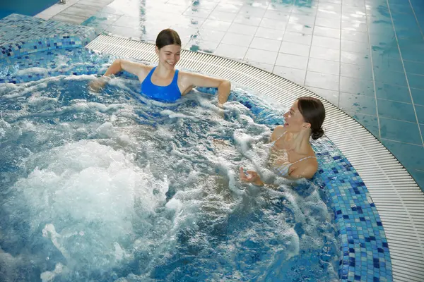 2人の若い女性がプールで熱い浴槽で休んでいます トレーニングクラスの後にリラックスするために泡風呂を取る水和療法治療を楽しむ女性スポーツウーマン — ストック写真