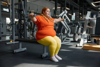 Kol kası pompalayan halterler kullanan şişman bir kadının portresi. Obez kız spor salonunda çalışıyor. Diyet ve zindelik kavramı